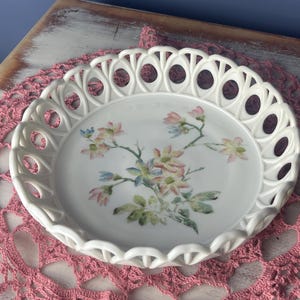 May include: White ceramic plate with a delicate openwork rim and a floral design featuring pink and white flowers. The plate is on a pink crocheted doily.