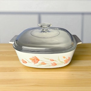 May include: A white casserole dish with a clear glass lid. The dish features a floral design in shades of orange and green. The handles are silver. The dish is on a wooden surface.