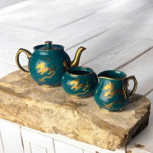 May include: A teal tea set with gold dragon accents. The set includes a teapot, a sugar bowl, and a creamer. The teapot has a curved handle and a lid with a gold knob. The sugar bowl is round, and the creamer has a curved handle.