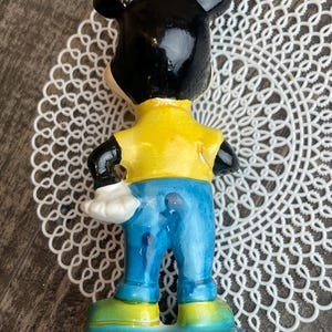 May include: A ceramic figurine of Mickey Mouse wearing a yellow shirt, blue jeans, and black shoes. The figurine is standing on a blue base.