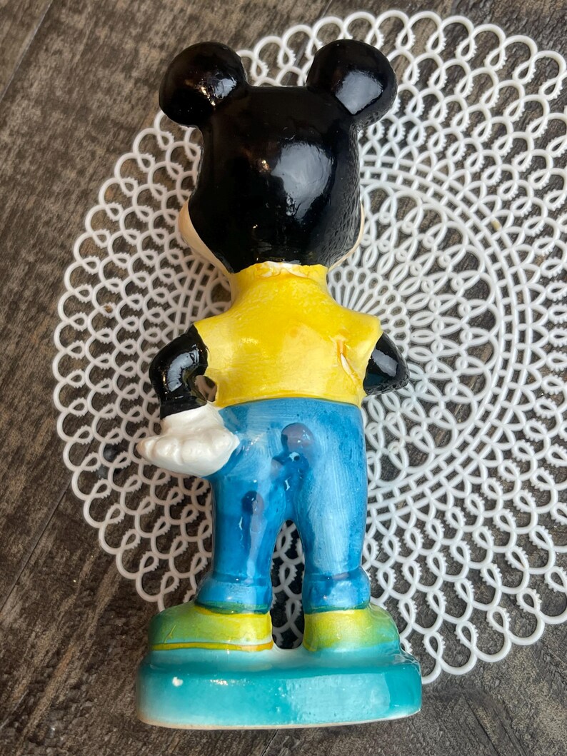 May include: A ceramic figurine of Mickey Mouse wearing a yellow shirt, blue jeans, and black shoes. The figurine is standing on a blue base.