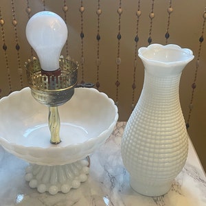 May include: Two white milk glass lamps, one with a bowl-shaped base and a light bulb, the other a vase-shaped lamp.