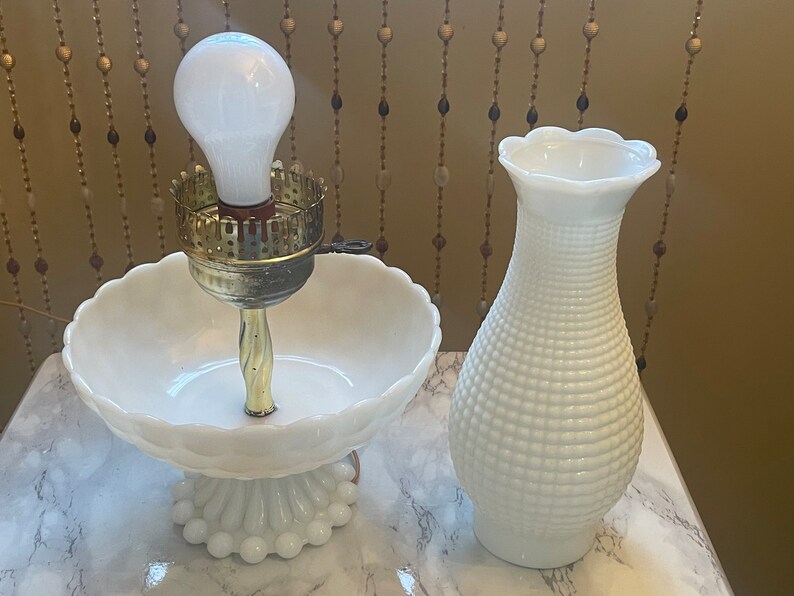 May include: Two white milk glass lamps, one with a bowl-shaped base and a light bulb, the other a vase-shaped lamp.