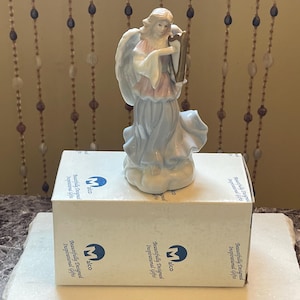 May include: A porcelain figurine of an angel playing a harp. The angel is wearing a blue and pink dress and has a halo. The figurine is sitting on a cloud base. The figurine is on a white box with the words "Beautifully Designed Inspirational Gifts" printed on it.
