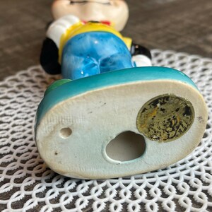 May include: A ceramic figurine of a cartoon character wearing a yellow shirt, blue pants, and a red bow tie. The figurine is standing on a blue base with a gold sticker that reads "Made in Japan".