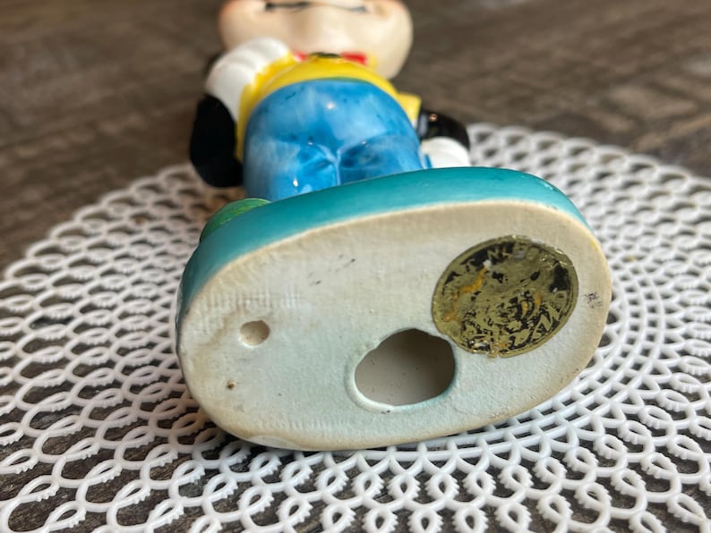 May include: A ceramic figurine of a cartoon character wearing a yellow shirt, blue pants, and a red bow tie. The figurine is standing on a blue base with a gold sticker that reads "Made in Japan".