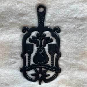 May include: Black cast iron trivet with a decorative design featuring a stylized flower, a star, and three utensils. The trivet has a handle for hanging.