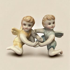 May include: A porcelain figurine depicting two cherubs in a playful embrace. One cherub wears a yellow outfit, the other a blue one. Both have blonde hair, small wings, and are kneeling. The figurine is a decorative item.