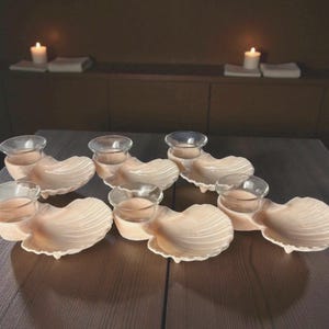 May include: Six seashell-shaped candle holders with clear glass votive cups. The holders are a light beige color and sit on a dark brown wooden table. Two lit candles are in the background.