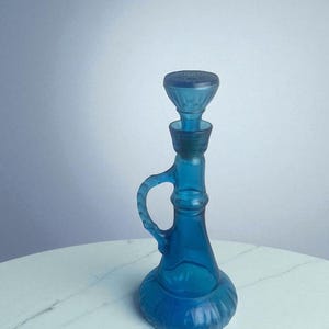 May include: A vintage blue glass decanter with a handle and stopper. The decanter has a bulbous base and a slender neck, with a decorative stopper. The handle is curved, and the glass has a textured appearance. The decanter is sitting on a white surface.