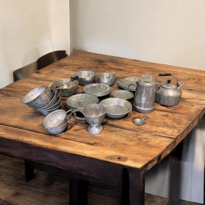 May include: A collection of vintage metal kitchenware, including cups, bowls, plates, a teapot, and a kettle, arranged on a rustic wooden table. The items are in various sizes and shapes, with a uniform silver color, suggesting a cohesive set.