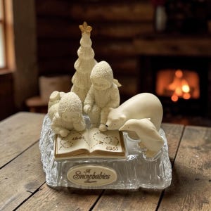 May include: A Snowbabies figurine featuring two snow babies, a polar bear, and a dolphin gathered around an open book. A Christmas tree with a gold star and a clear base complete the scene. The base reads "A Snowbabies Collection."
