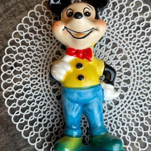 May include: A ceramic figurine of Mickey Mouse wearing a yellow shirt, blue pants, and a red bow tie. He is smiling and has a black nose and ears.