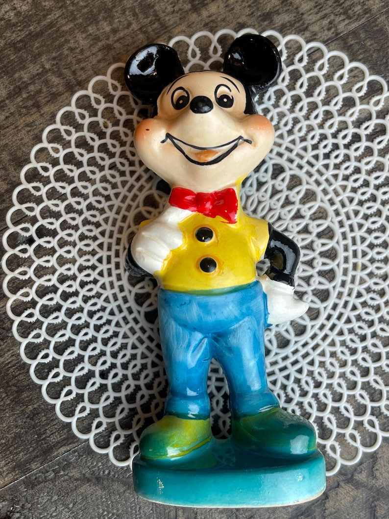 May include: A ceramic figurine of Mickey Mouse wearing a yellow shirt, blue pants, and a red bow tie. He is smiling and has a black nose and ears.