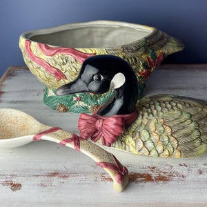May include: A ceramic goose-shaped serving bowl with a red and green floral design. The bowl is decorated with a red ribbon and a matching ladle.
