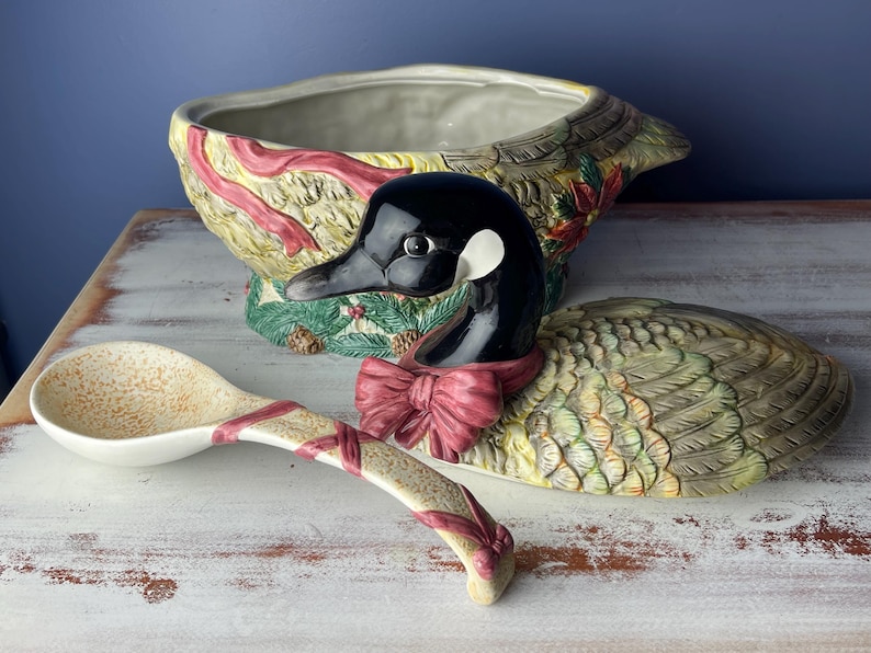 May include: A ceramic goose-shaped serving bowl with a red and green floral design. The bowl is decorated with a red ribbon and a matching ladle.
