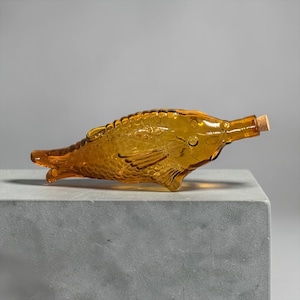 May include: A translucent amber-colored glass bottle shaped like a fish, featuring molded scale details along the body and fins. The bottle is positioned horizontally with a small cork stopper inserted into the narrow opening at the fish's mouth.