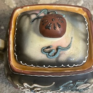 Op de afbeelding: Een vierkante theepot van keramiek met een bruin deksel en ingewikkelde bloem- en rolpatronen in tinten blauw, wit en goud. De theepot heeft een bruine glazuur en een witte rand.