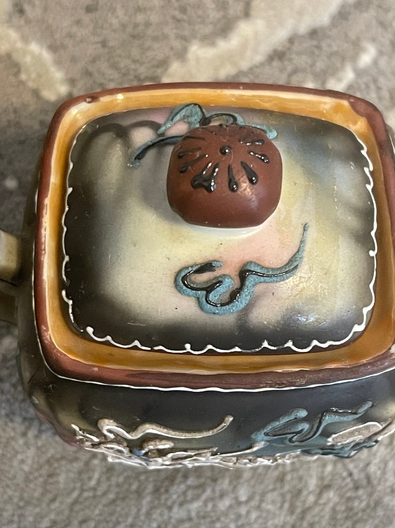 Op de afbeelding: Een vierkante theepot van keramiek met een bruin deksel en ingewikkelde bloem- en rolpatronen in tinten blauw, wit en goud. De theepot heeft een bruine glazuur en een witte rand.