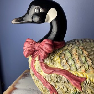 May include: A ceramic goose figurine with a red bow, detailed feathers, and a poinsettia flower. The goose is painted in shades of brown, green, and yellow.