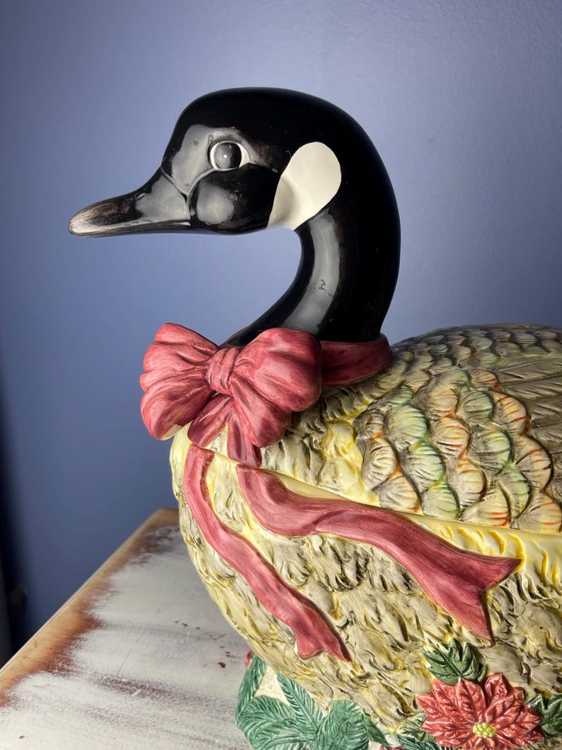 May include: A ceramic goose figurine with a red bow, detailed feathers, and a poinsettia flower. The goose is painted in shades of brown, green, and yellow.