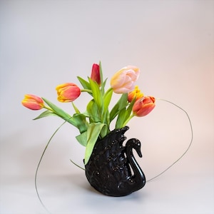 May include: A black swan-shaped vase holds a bouquet of colorful tulips. The tulips display shades of yellow, orange, and pink, with green stems and leaves. The vase is glossy and sits on a light gray surface.