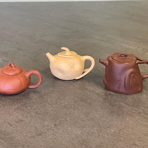 May include: Three small ceramic teapots in different colors and styles. The teapots are brown, beige, and reddish-brown.