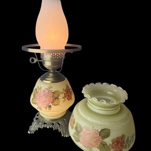 Puede incluir: L&aacute;mpara de mesa vintage con dise&ntilde;o floral. La l&aacute;mpara presenta una base y pantalla de color crema adornadas con rosas rosas y rojas. La l&aacute;mpara tiene una base de metal y una chimenea de vidrio esmerilado.