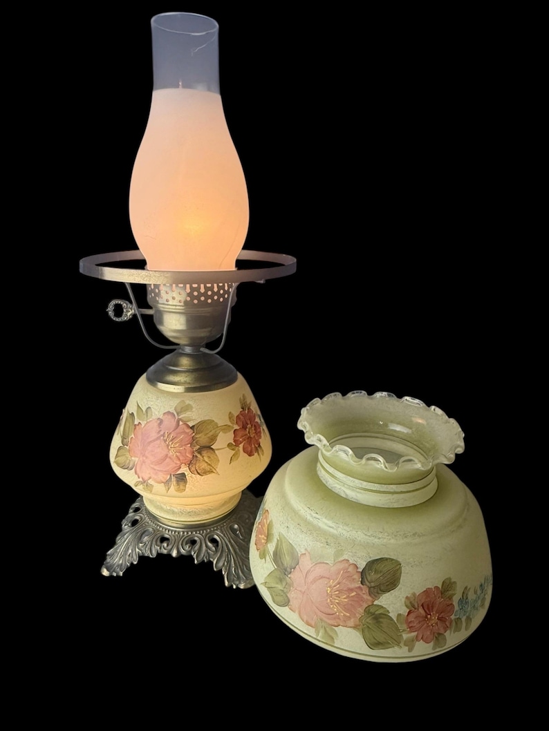 Puede incluir: L&aacute;mpara de mesa vintage con dise&ntilde;o floral. La l&aacute;mpara presenta una base y pantalla de color crema adornadas con rosas rosas y rojas. La l&aacute;mpara tiene una base de metal y una chimenea de vidrio esmerilado.