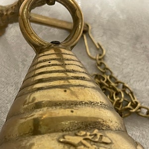 May include: A brass bell with a decorative design and a chain attached to a hook. The bell is hanging from a brass rod.