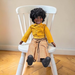 May include: A ventriloquist doll with dark skin, curly black hair, and glasses. The doll wears a long-sleeved mustard yellow button-down shirt and tan trousers. It has a wide, open-mouthed expression and is wearing black shoes on its feet.