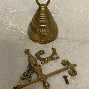 May include: A brass bell with a decorative design and a hanging mechanism. The bell has a loop at the top for hanging and a detailed design on the side. The hanging mechanism is made of brass and has a chain and a hook.
