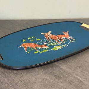 May include: Oval serving tray with a teal blue surface and a black border. The tray features a deer illustration in shades of brown and green. The handles are wrapped in a light-colored woven material.