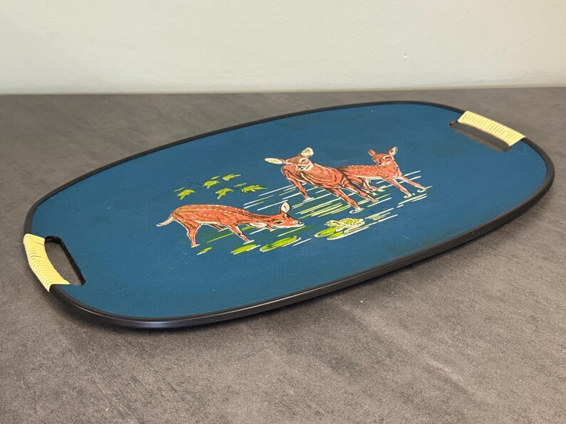 May include: Oval serving tray with a teal blue surface and a black border. The tray features a deer illustration in shades of brown and green. The handles are wrapped in a light-colored woven material.