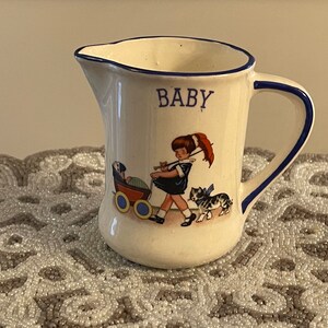 May include: A white ceramic creamer with a blue rim and handle. It features a colorful illustration of a girl pushing a baby carriage with a cat walking beside her. The word "BABY" is printed in blue on the front.
