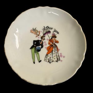 May include: A white, scalloped-edge plate with a gold rim. The plate features a romantic illustration of a couple under an umbrella, with the words "I love you" and a cupid. Pink hearts are scattered around the figures.