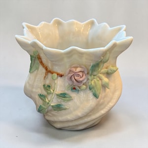 May include: A cream-colored porcelain vase with a scalloped rim and a shell-like texture. The exterior is decorated with a three-dimensional ceramic rose in soft pink, accompanied by green leaves and a brown branch accent attached to the side.