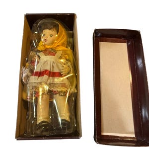 May include: A vintage doll in a box. The doll has a yellow headscarf, a white apron with a red stripe, and a floral patterned dress. The doll is in a brown cardboard box with a clear plastic cover.