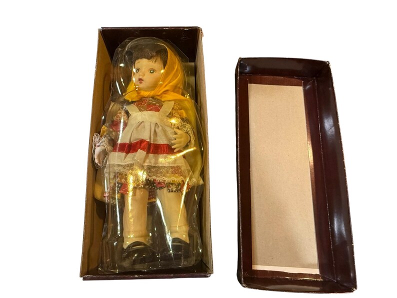 May include: A vintage doll in a box. The doll has a yellow headscarf, a white apron with a red stripe, and a floral patterned dress. The doll is in a brown cardboard box with a clear plastic cover.