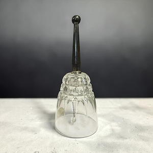 May include: A clear glass bell with a faceted design and a dark, elongated handle. The bell has a silver-colored metal base and a small clapper inside. The handle is topped with a small, round knob.