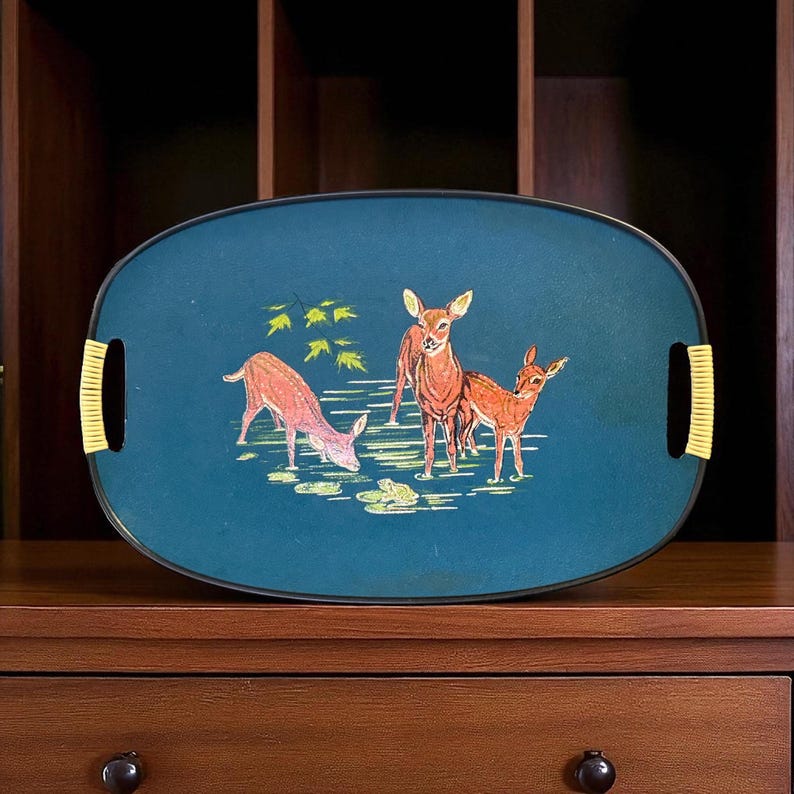 May include: Oval serving tray with a teal background and a deer scene. The tray features three deer in shades of brown and pink, with yellow accents. The handles are yellow. A decorative piece for serving.