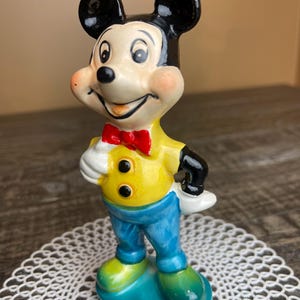 May include: A ceramic figurine of Mickey Mouse wearing a yellow shirt with black buttons, a red bow tie, and blue pants. He is standing on a blue base with green accents.