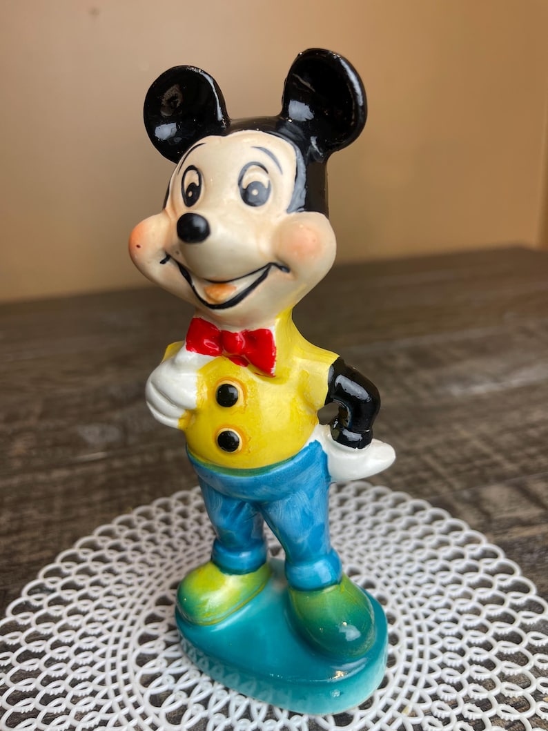 May include: A ceramic figurine of Mickey Mouse wearing a yellow shirt with black buttons, a red bow tie, and blue pants. He is standing on a blue base with green accents.