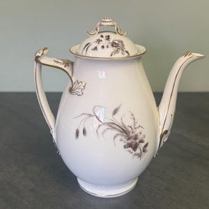 May include: White porcelain teapot with a lid, featuring floral and botanical designs in brown and purple. The teapot has a curved spout, a handle with gold accents, and gold trim. A decorative piece for serving tea.