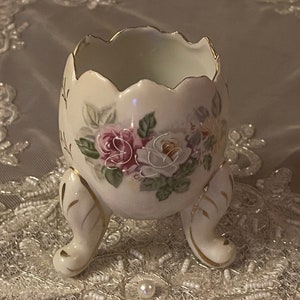 May include: A white porcelain egg-shaped vase with three legs and a floral design featuring pink and white roses. The vase has a gold trim around the top edge.