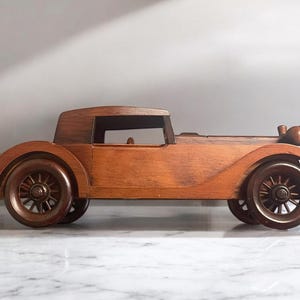 May include: A wooden model of a vintage car, featuring a classic design with a closed top and spoked wheels. The car is crafted from a warm-toned wood, showcasing the grain. The model is a decorative item.