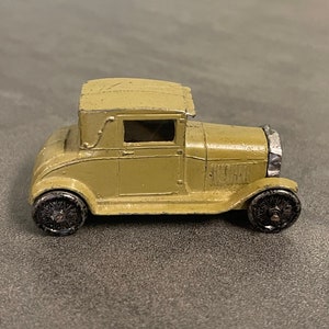May include: A vintage toy car made of metal, painted in a light brown color. The car has a closed roof and a detailed front grille. The car has black rubber tires.
