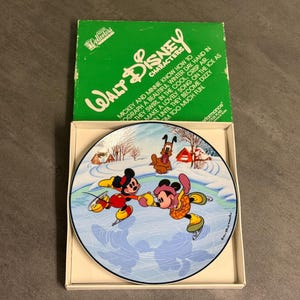 May include: A Walt Disney Characters collector's plate featuring Mickey and Minnie Mouse ice skating on a frozen pond. The plate is in a box with a green lid that reads "Walt Disney Characters". The plate has a black border.