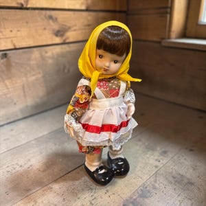 May include: A vintage doll with a yellow headscarf and a colorful patchwork dress. The doll has a white apron with red trim and black shoes. The doll is standing on a wooden surface.