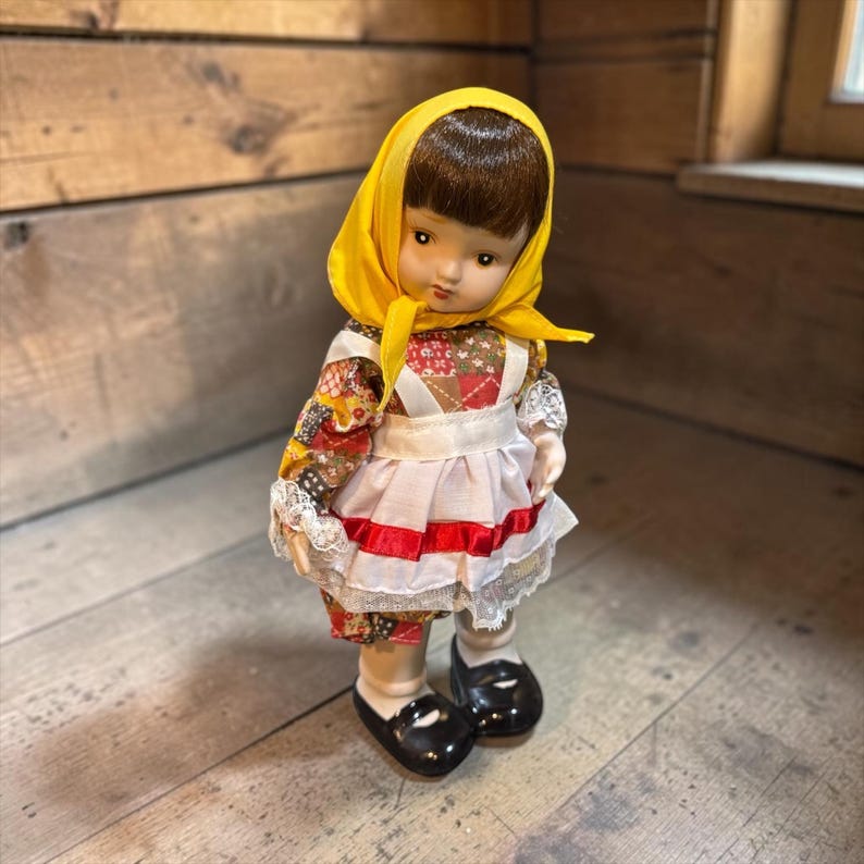 May include: A vintage doll with a yellow headscarf and a colorful patchwork dress. The doll has a white apron with red trim and black shoes. The doll is standing on a wooden surface.